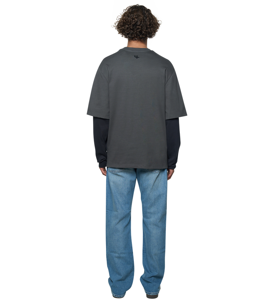 Person wearing a dark gray shirt over a black long-sleeve shirt with blue jeans on a white background